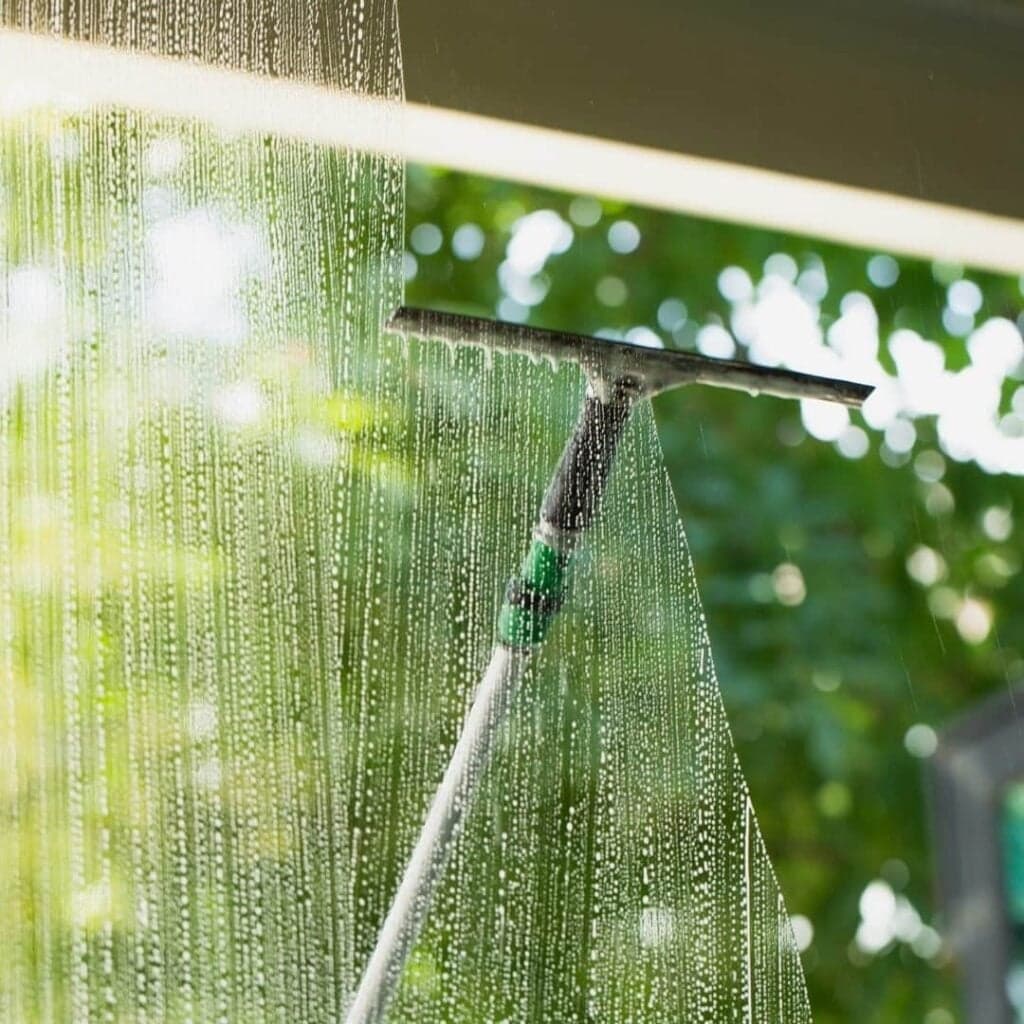 A close-up of a squeegee on a long pole being used to clean a large window, leaving soapy water streaks. The background is a bright, blurry view of green trees and foliage.