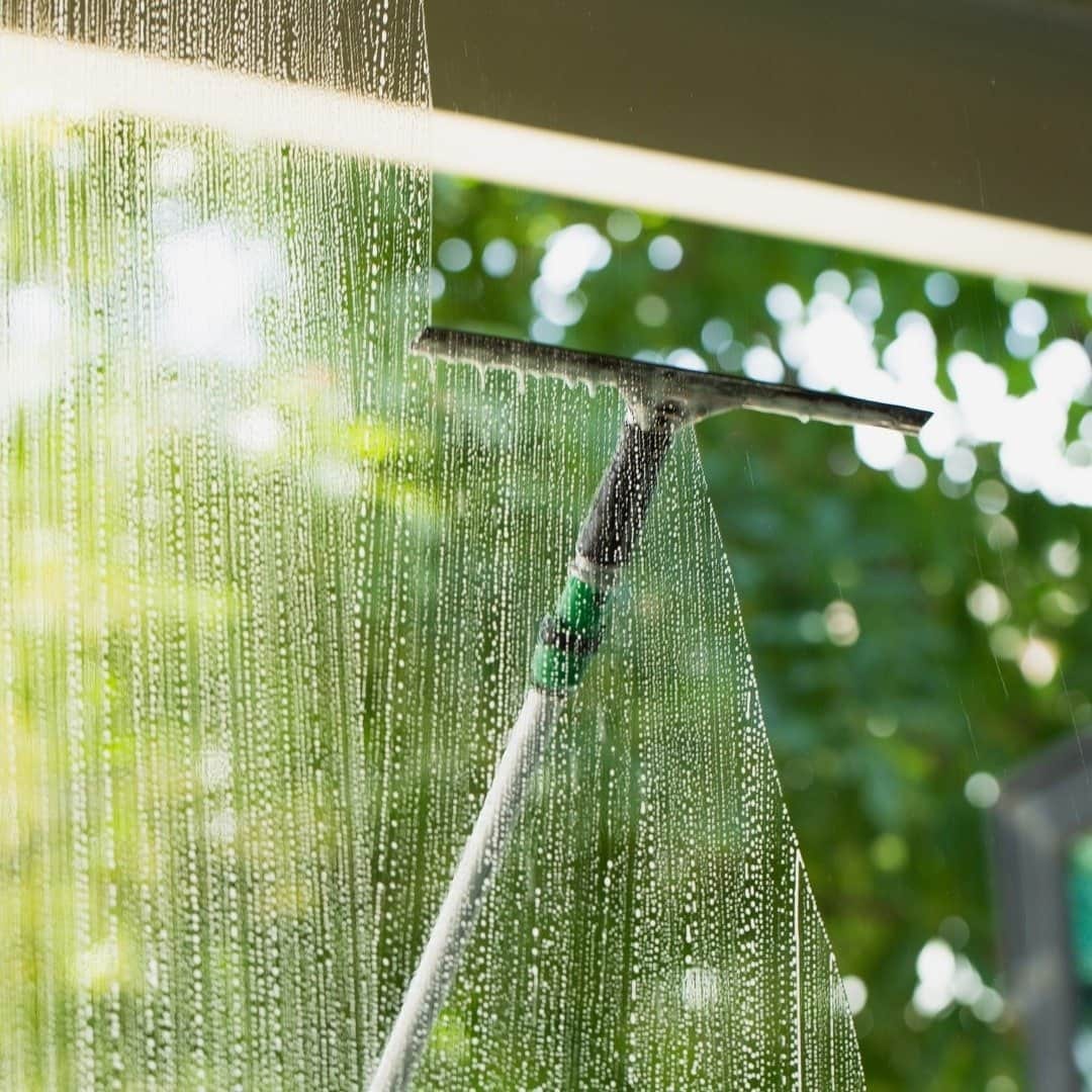 A close-up of a squeegee on a long pole being used to clean a large window, leaving soapy water streaks. The background is a bright, blurry view of green trees and foliage.