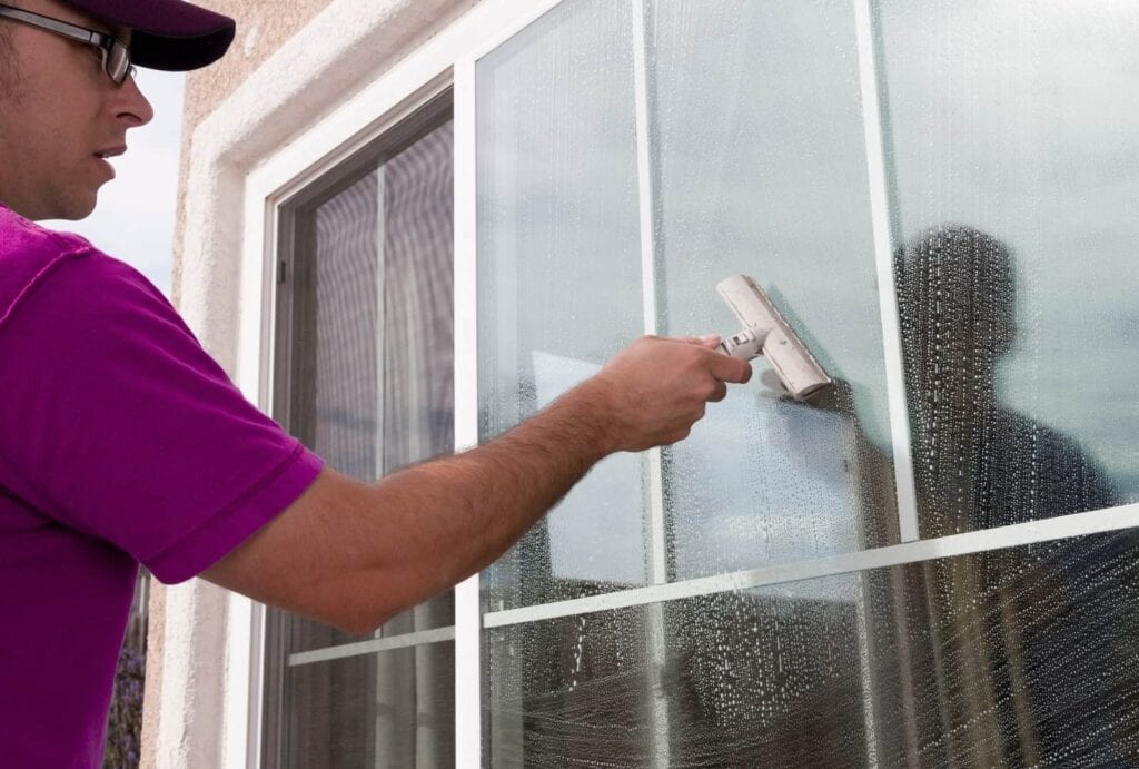 A close-up of a worker in a purple/pink shirt, baseball cap, and glasses, using a blue and white fabric scrubber to apply soapy water to a glass window with white trim. The background is a sunny yard with plants.