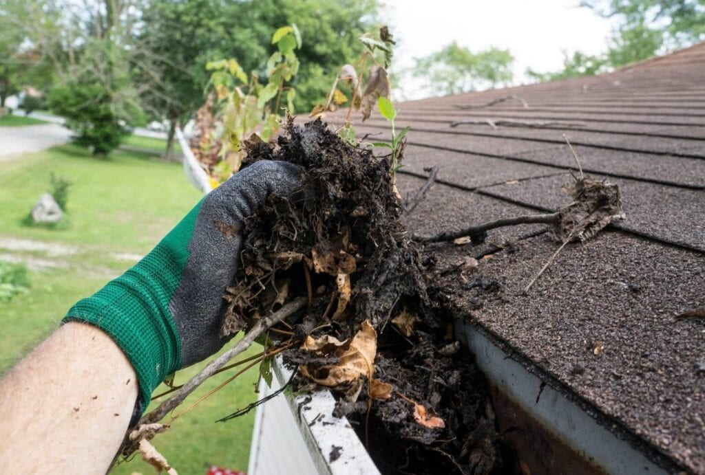 A hand wearing a green and black work glove is reaching into a roof gutter to pull out a large clump of wet, dark debris, including dirt, leaves, and small branches. The gutter is attached to a shingled roof with a grassy residential area in the background.
