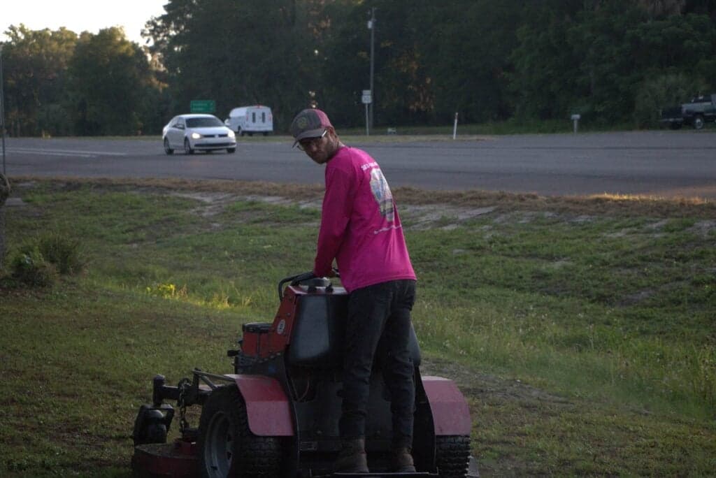 A man in a bright pink long-sleeve shirt and a baseball cap is operating a stand-on lawnmower in a large, overgrown grassy area next to a paved road. A white car and a trailer are visible on the road in the distance.