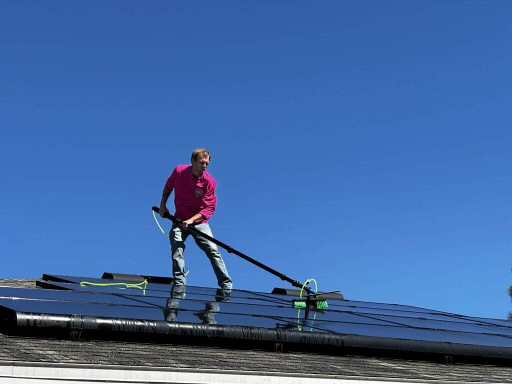 A man in a pink shirt and jeans is standing on a dark, reflective rooftop covered in solar panels, using a long pole with a brush attachment to clean them under a bright blue sky.
