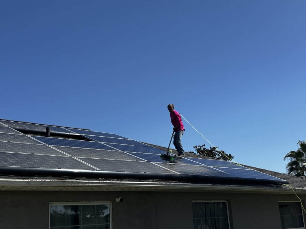 A worker in a pink long-sleeve shirt is using a long brush on a pole, connected to a hose, to clean dark, reflective solar panels installed on a residential roof under a bright blue sky. The background shows the house's siding and window.