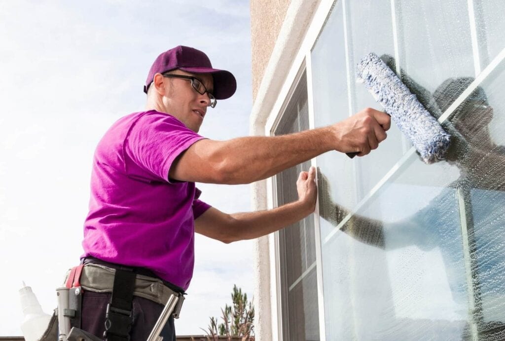 A worker in a pink t-shirt, dark cap, and glasses is pouring cleaning solution from a white bottle onto a window cleaning tool with a blue and white fabric head. He is cleaning a large glass window of a house on a sunny day.