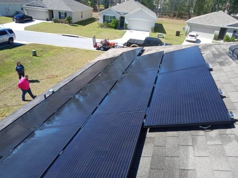 An aerial view of a residential roof covered in dark shingles with a large array of reflective black solar panels. Two workers, one in a pink shirt, are visible on the left edge of the roof. The background shows suburban houses, driveways, and a trailer attached to a truck.