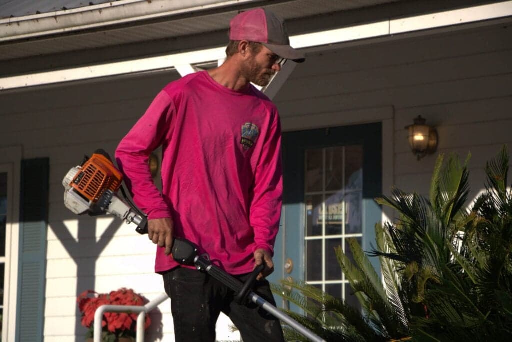 A professional landscaper in a pink long-sleeve shirt and grey baseball cap using a gas-powered string trimmer to edge a residential yard near a house.