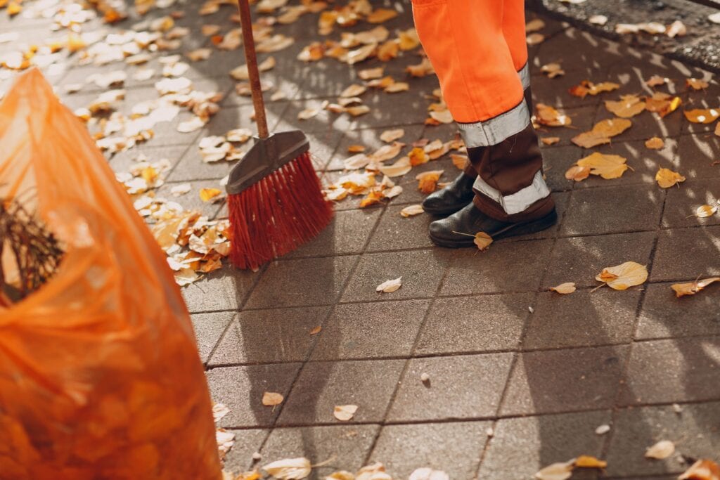 A street cleaner in high-visibility orange trousers and reflective boots sweeping dry autumn leaves on a paved sidewalk with a red broom.