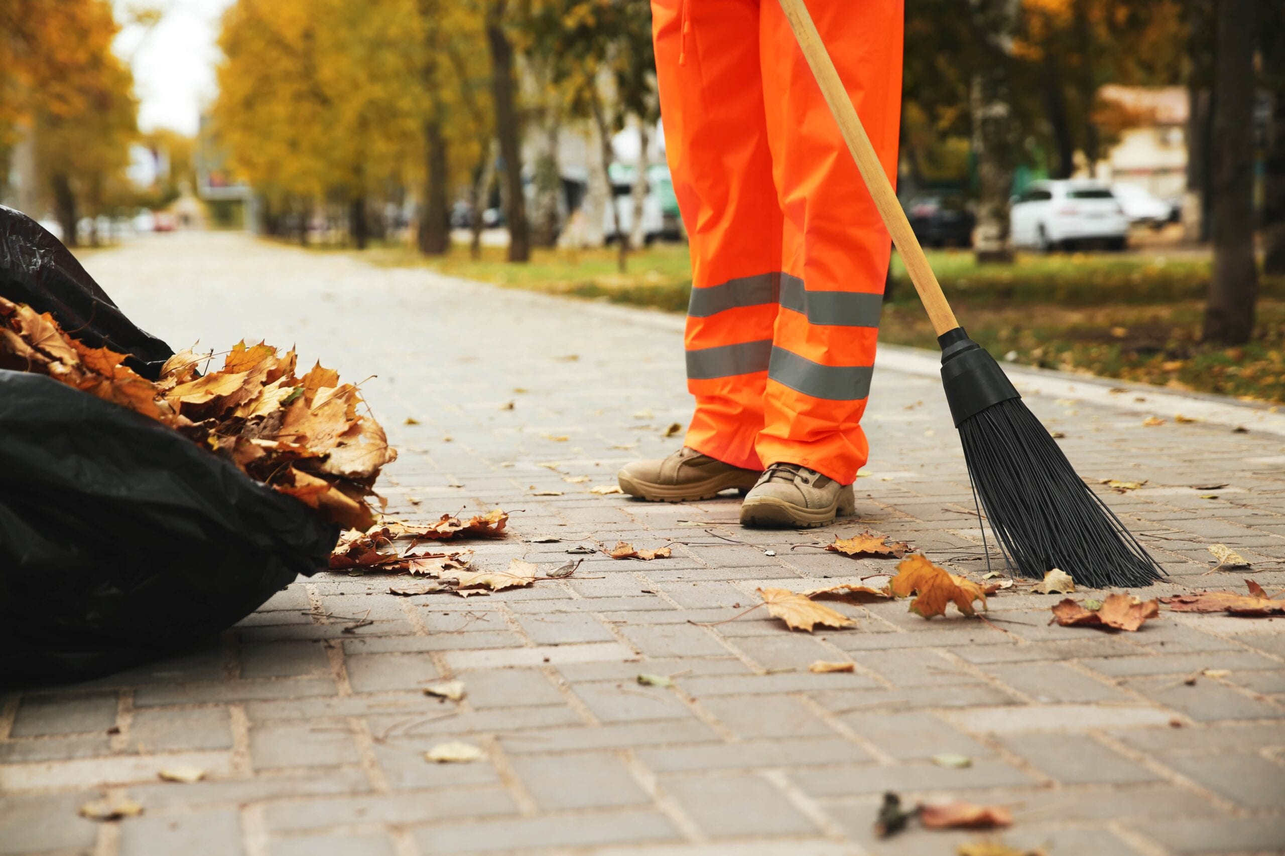 A street cleaner in bright orange high-visibility safety gear using a large broom to sweep fallen autumn leaves into a black waste bag on a paved park walkway.