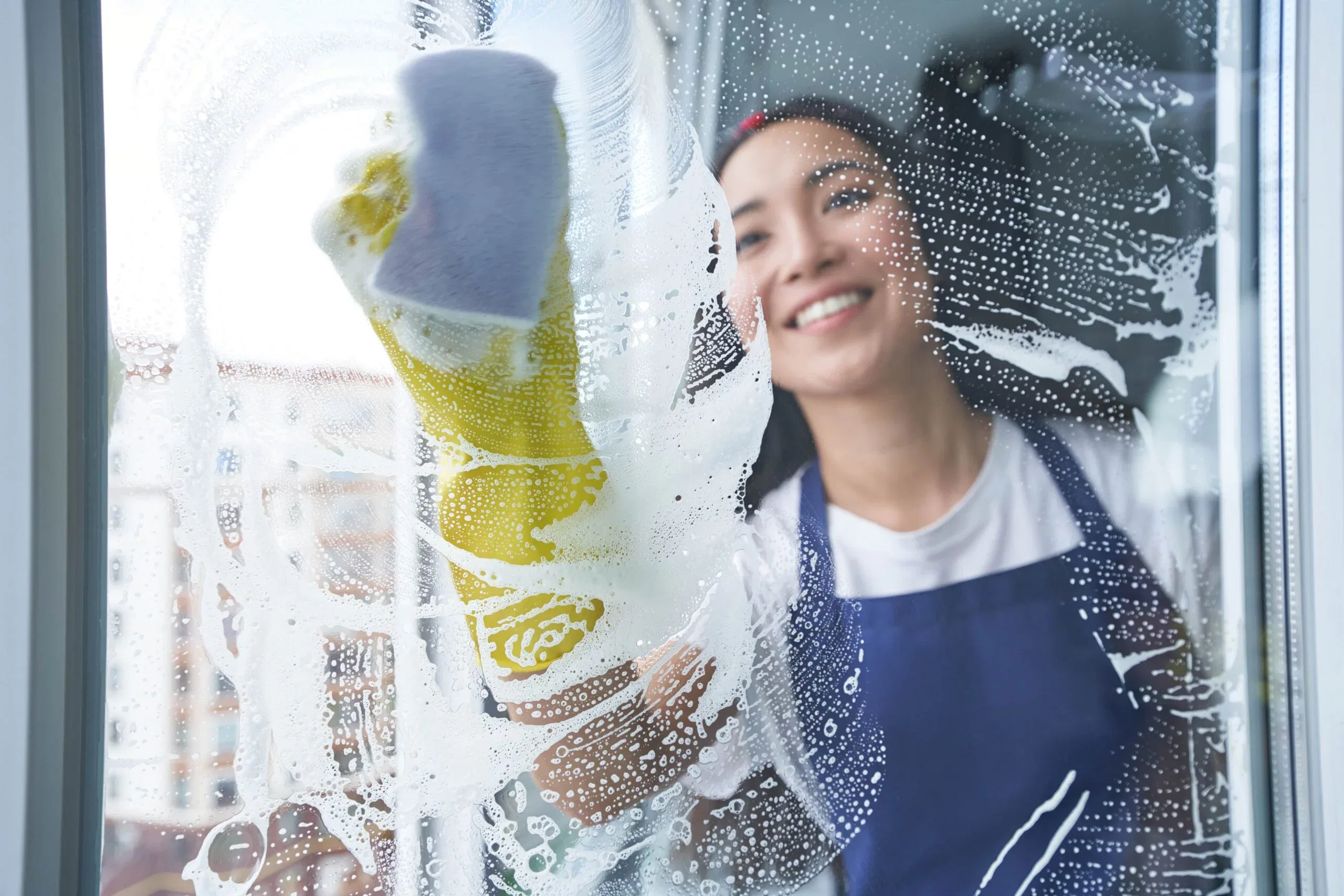 A smiling young woman wearing yellow rubber gloves and a blue apron uses a sponge to scrub soapy lather onto a glass window pane from the outside.