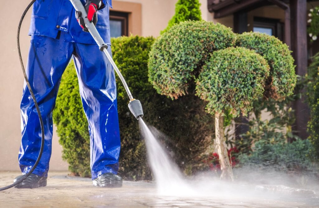A professional cleaner in blue waterproof gear using a high-pressure washer to clean a stone paver driveway in front of residential landscaping.