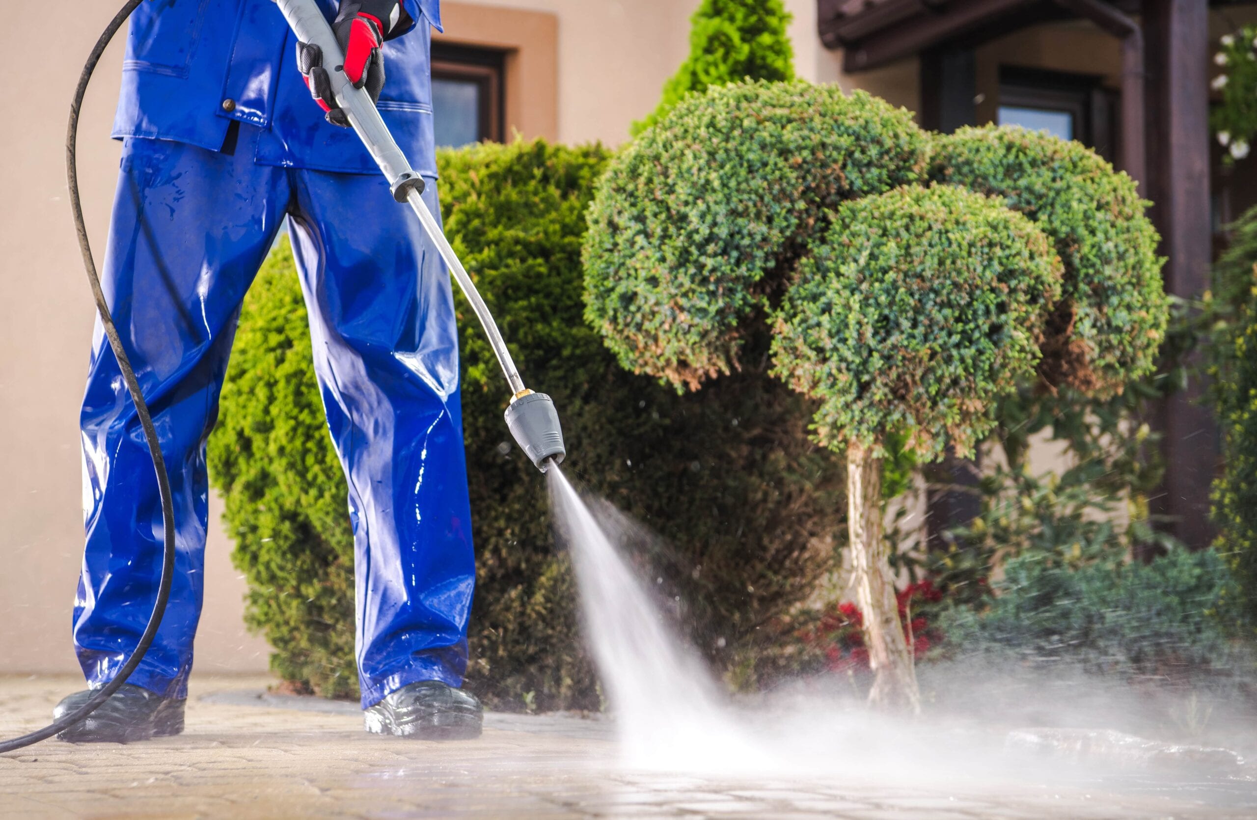 A professional cleaner in blue waterproof gear using a high-pressure washer to clean a stone paver driveway in front of residential landscaping.