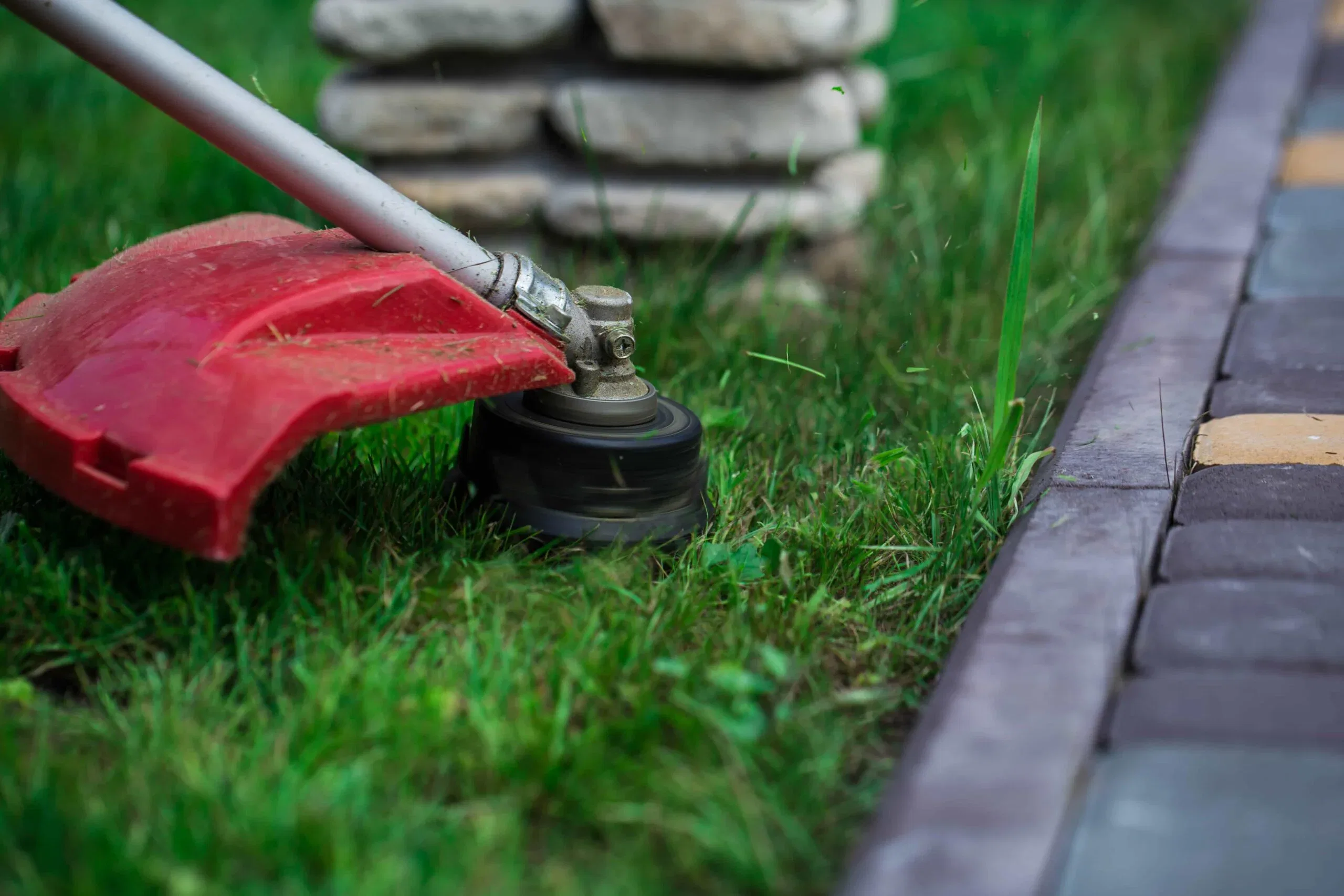 A red string trimmer head cutting grass along a brick walkway and stone garden border, providing professional lawn edging service in The Villages, Florida.
