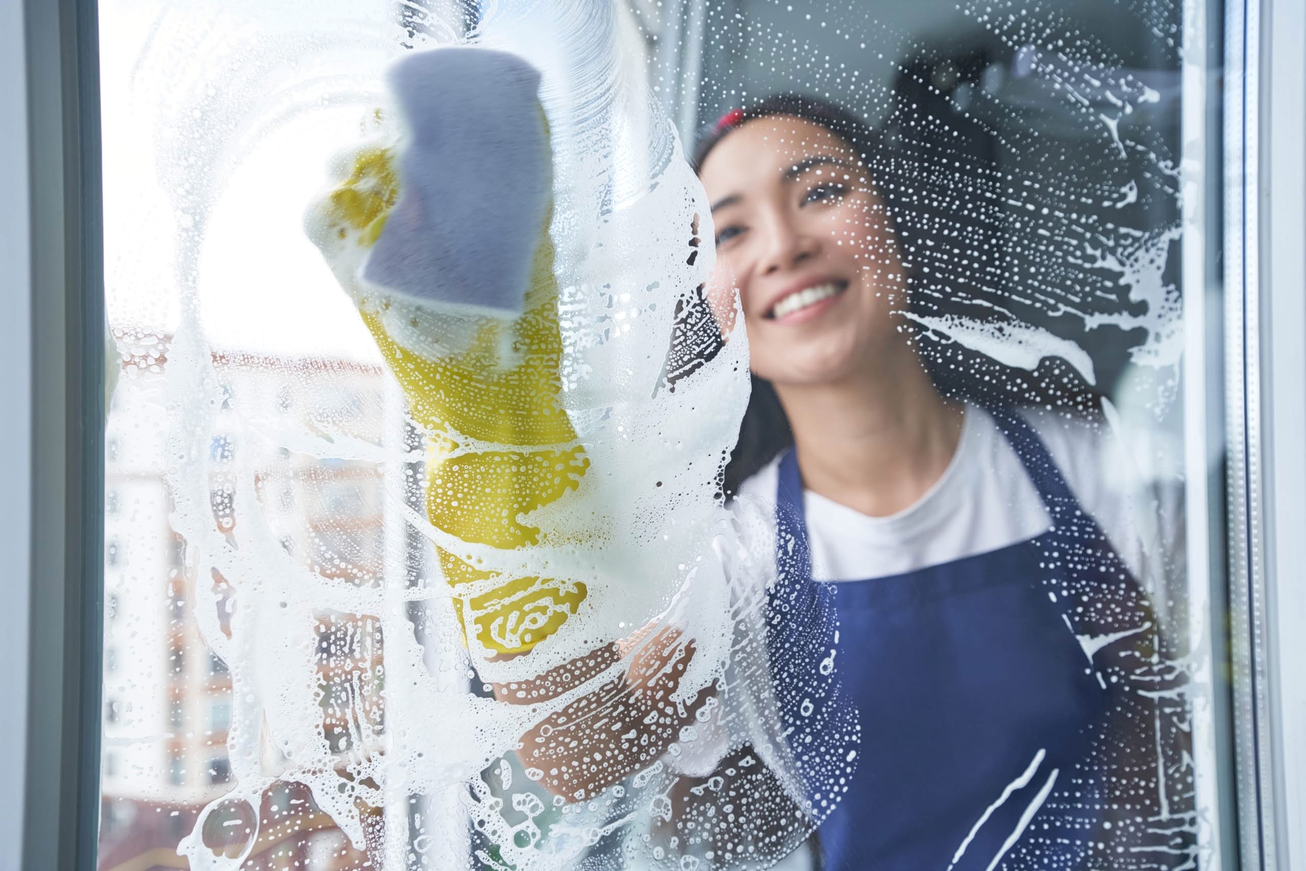 A smiling young woman wearing yellow rubber gloves and a blue apron uses a sponge to scrub soapy lather onto a glass window pane from the outside.